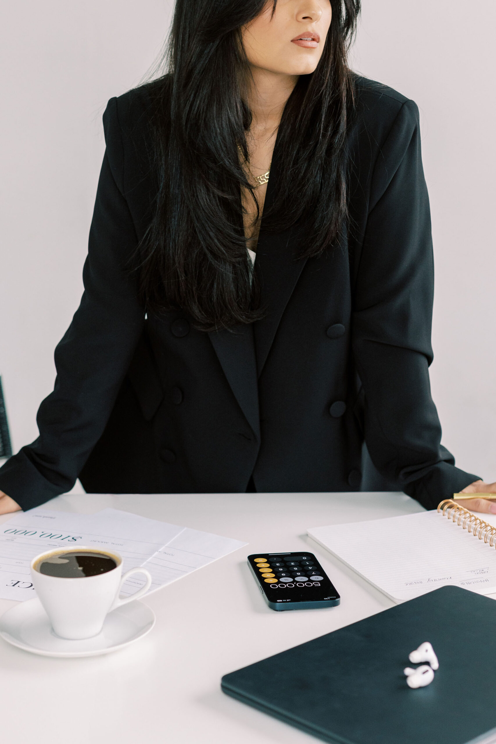 A woman wearing a black blazer, leaning against a desk, deep in thought. On the desk there is a cup of black coffee, calculator, and apple airpods