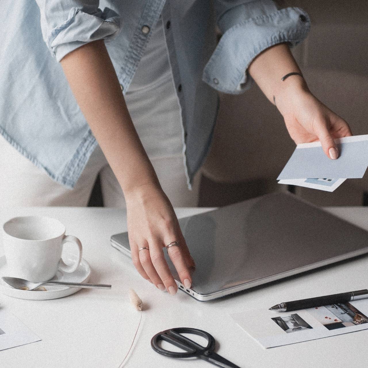 faceless woman in blue long sleeve polo holding laptop and blue card and images, with a mug and spoon on the side, scissors, and photos